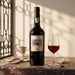 A bottle of Duna Antonia 10 year old tawny port stands on a rustic stone surface beside a filled port glass, with a small metal cup, cloth, and decorative stones in a warm sunlit setting.