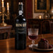 A bottle of BIN 27 Reserve Port by Fonseca sits on a wooden table beside a glass of deep port wine and a chocolate dessert with cherries, set in a warmly lit, elegant interior.
