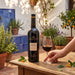 A bottle of Gonzalez Byass Nectar Pedro Ximenez Sherry stands on a wooden table beside a hand holding a glass of sherry, with potted plants and a tiled wall in the background.