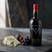 A bottle of Six Grapes Graham's Reserve Port wine stands on a dark stone surface next to a wedge of blue cheese, figs, and a cluster of dark grapes. The bottle has a red cap and a black label with white and gold text.