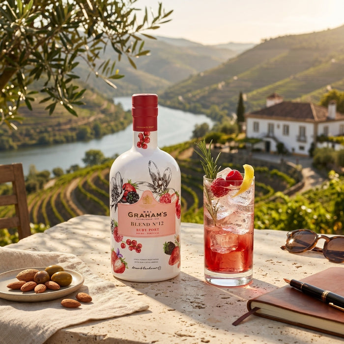Graham's Blend No.12 Port wine bottle with a glass of red wine and garnishes on a scenic outdoor table.