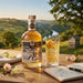 A bottle of Hive Mind Traditional Mead and a filled glass of the golden beverage are arranged on a wooden table outdoors, with a scenic countryside landscape in the background and small flowers and an open notebook in the foreground.