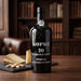 A dark glass bottle of Kopke 20 Years Old Tawny Port stands on a wooden table in a warm, book-filled room. A cheese and almond garnish sits on a slate