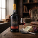 A bottle of Sandeman Founders Reserve red wine stands on a rustic wooden table beside a filled wine glass, a wedge of blue cheese, grapes, and a knife, with a softly blurred indoor room in the background.