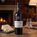 A bottle of Taylor's Late Bottled Vintage Port wine stands on a wooden table in a warm, cozy room setting with a wine glass and cheese on a slate board nearby.