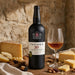 A bottle of Taylor's 30 Year Old Tawny Port stands on a wooden table beside a filled wine glass, surrounded by cheese wedges, pear slices, walnuts, and a cork, against a rustic stone wall background.