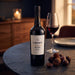 A bottle of Croft Vintage Port 2017 wine stands on a dark stone table beside a filled wine glass, with chocolate pieces nearby in a warmly lit indoor setting.