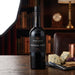 A bottle of FONSECA PORT Vintage Port 2016 stands on a wooden table in a warm, dimly lit room with bookshelves, a chair, and cheese and crackers on a slate board nearby.