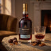 A bottle of Graham's 40 Year Old port stands on a stone table beside a filled rocks glass and several chocolate-covered sweets, with a softly blurred indoor setting in the background.