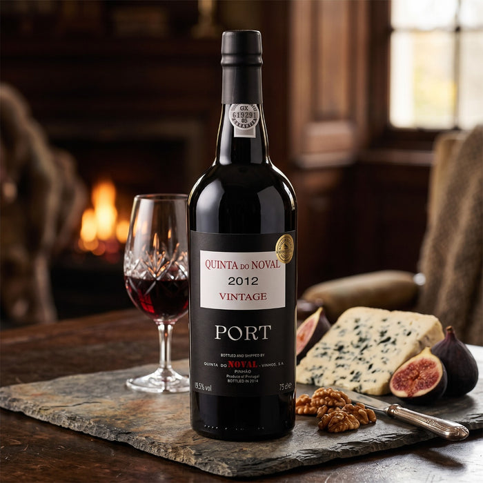 A bottle of Quinta do Noval 2012 Port wine stands on a stone serving board with a glass of port, cheese, figs, walnuts, and a knife in a warm, rustic dining setting.