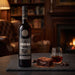 A bottle of Sandeman 20YO PX Sherry with chocolate garnish, displayed on a slate board next to a glass of the amber liquid, a chocolate brownie, and chocolate pieces, set against a warm, blurred background of a cozy living room with a fireplace.