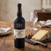 A bottle of Taylor's 20 Year Old Tawny Port wine stands on a rustic wooden table, styled with a wedge of yellow cheese, a cheese knife, and a wine glass in the background.